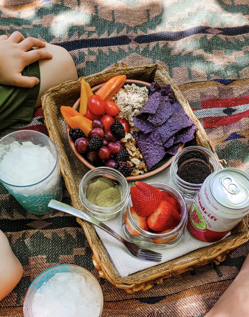 Picnic meal on picnic blanket. Tray contains peppers, blue corn chips, strawberries, sparkling water, grapes, and nuts.